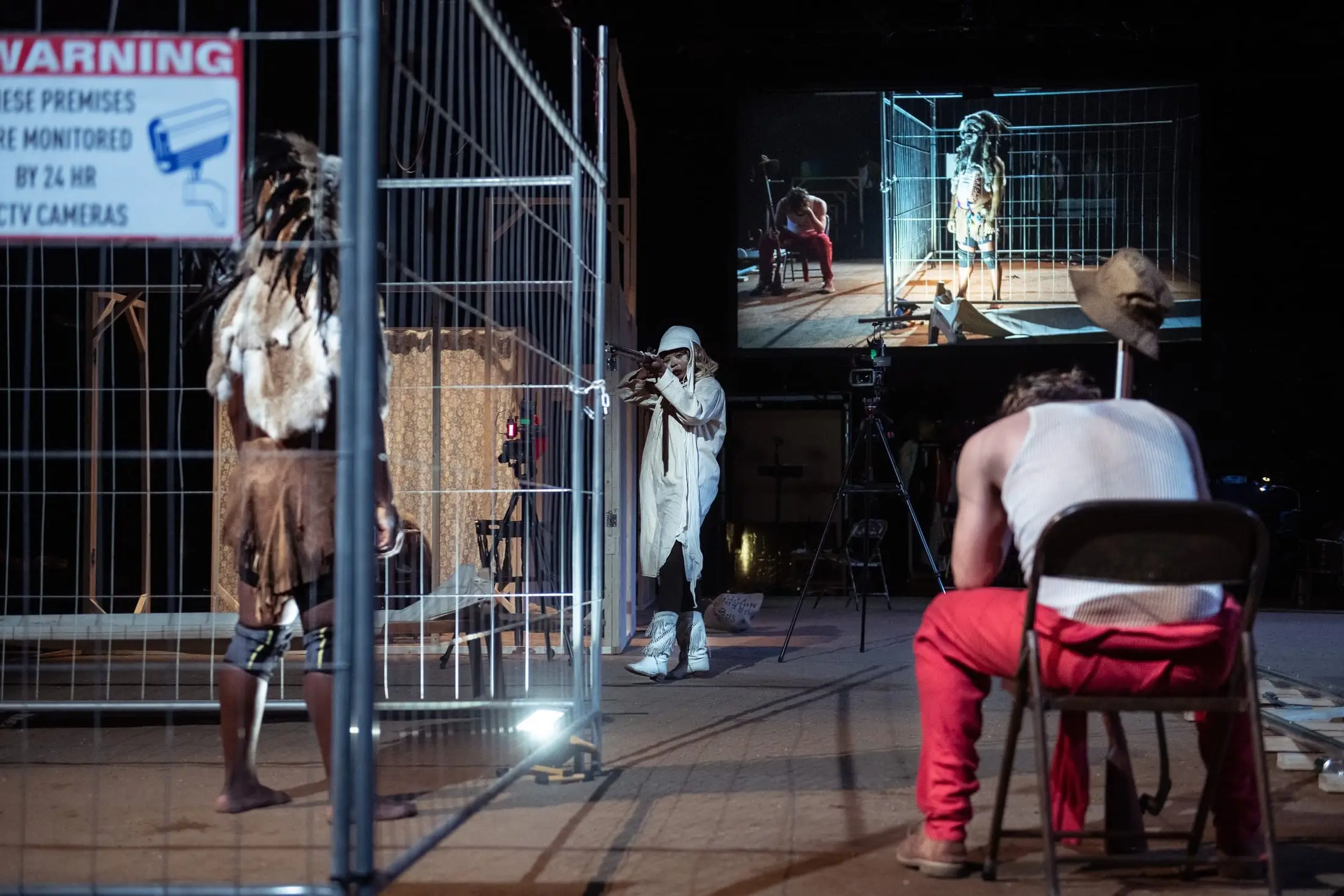 Performers behind a wire fence under a CCTV warning sign, a camera filming