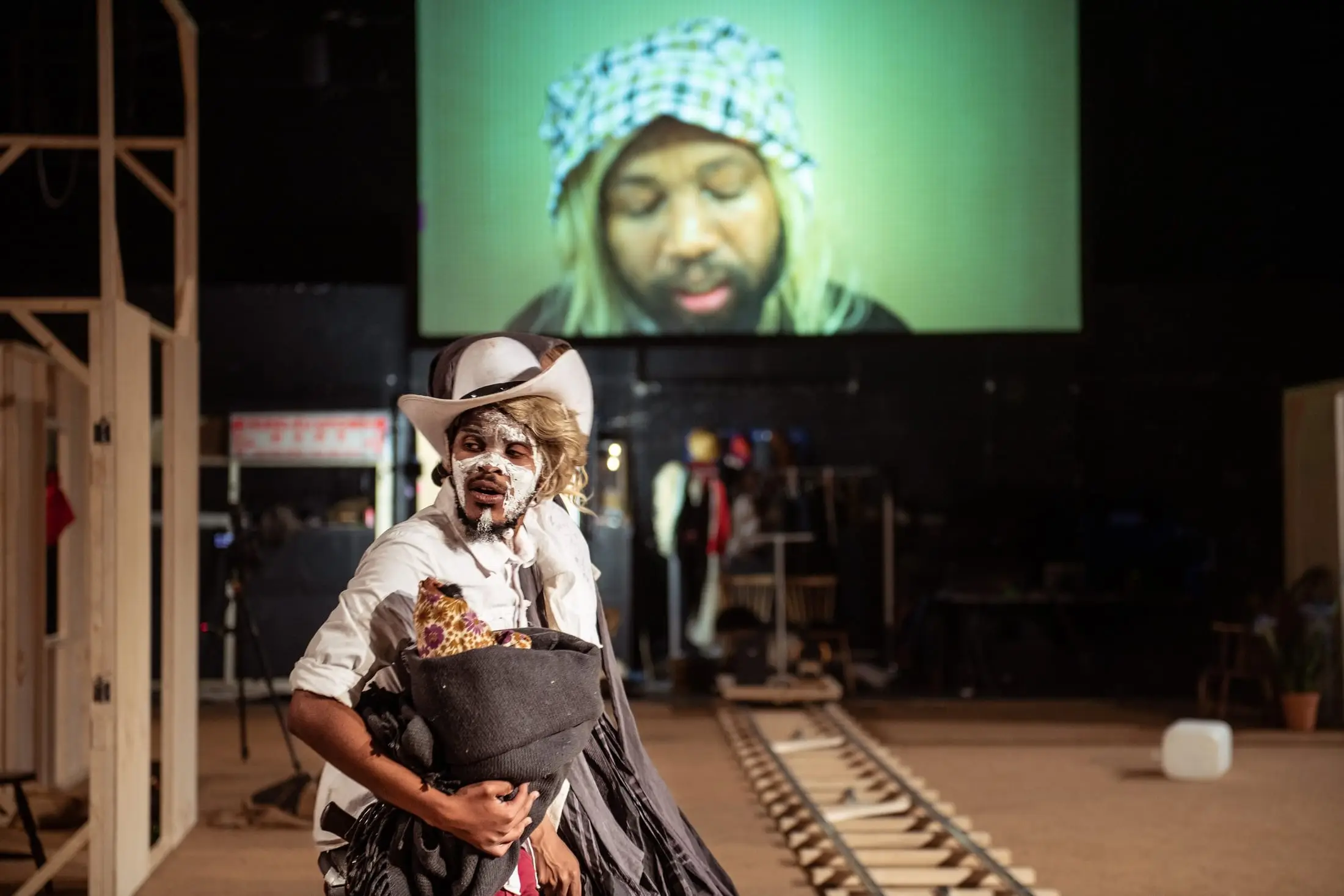 A performer in a cowboy hat clutches a bag, the projected face of an actor behind