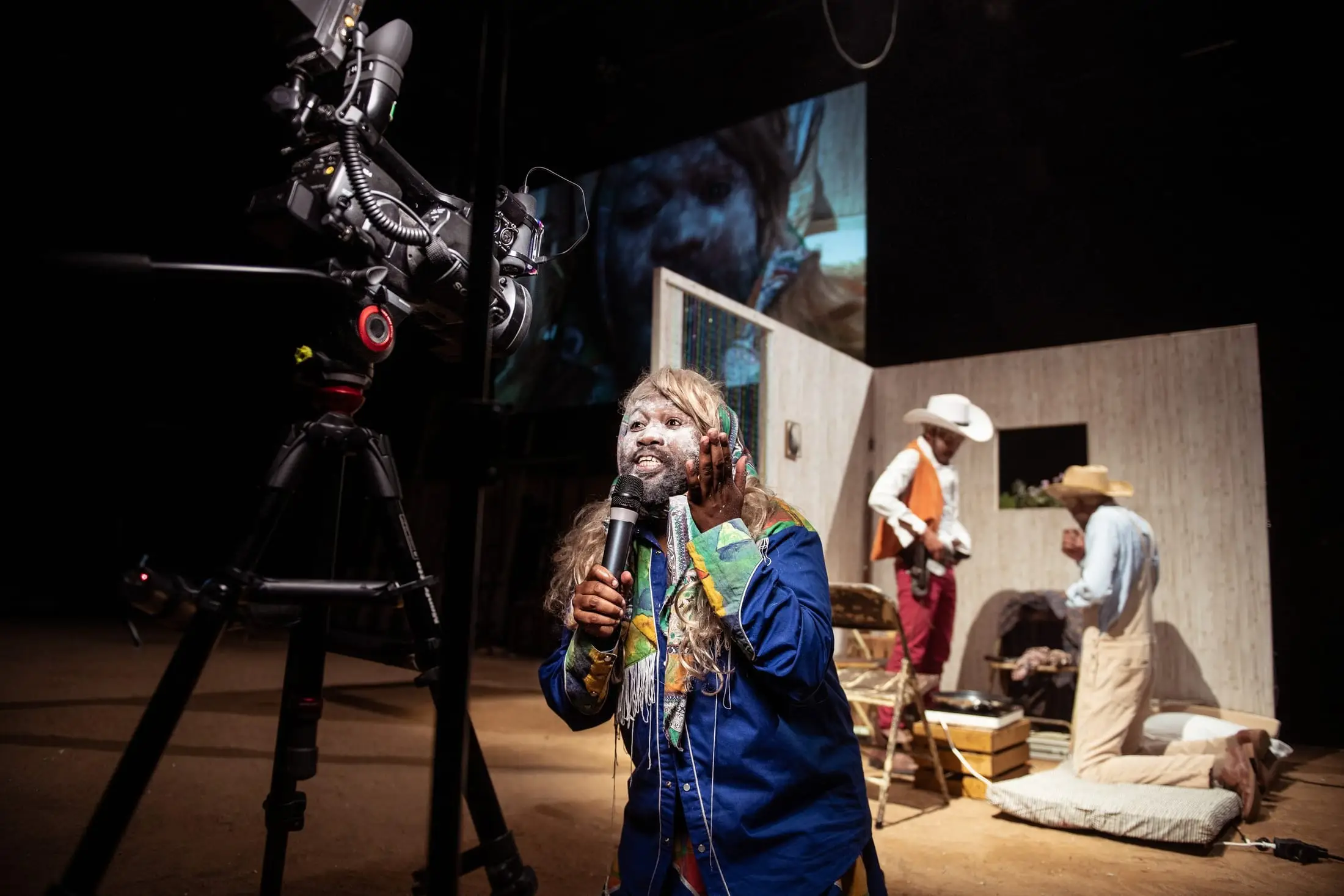 A performer sings into a microphone beside a camera crane, the town behind