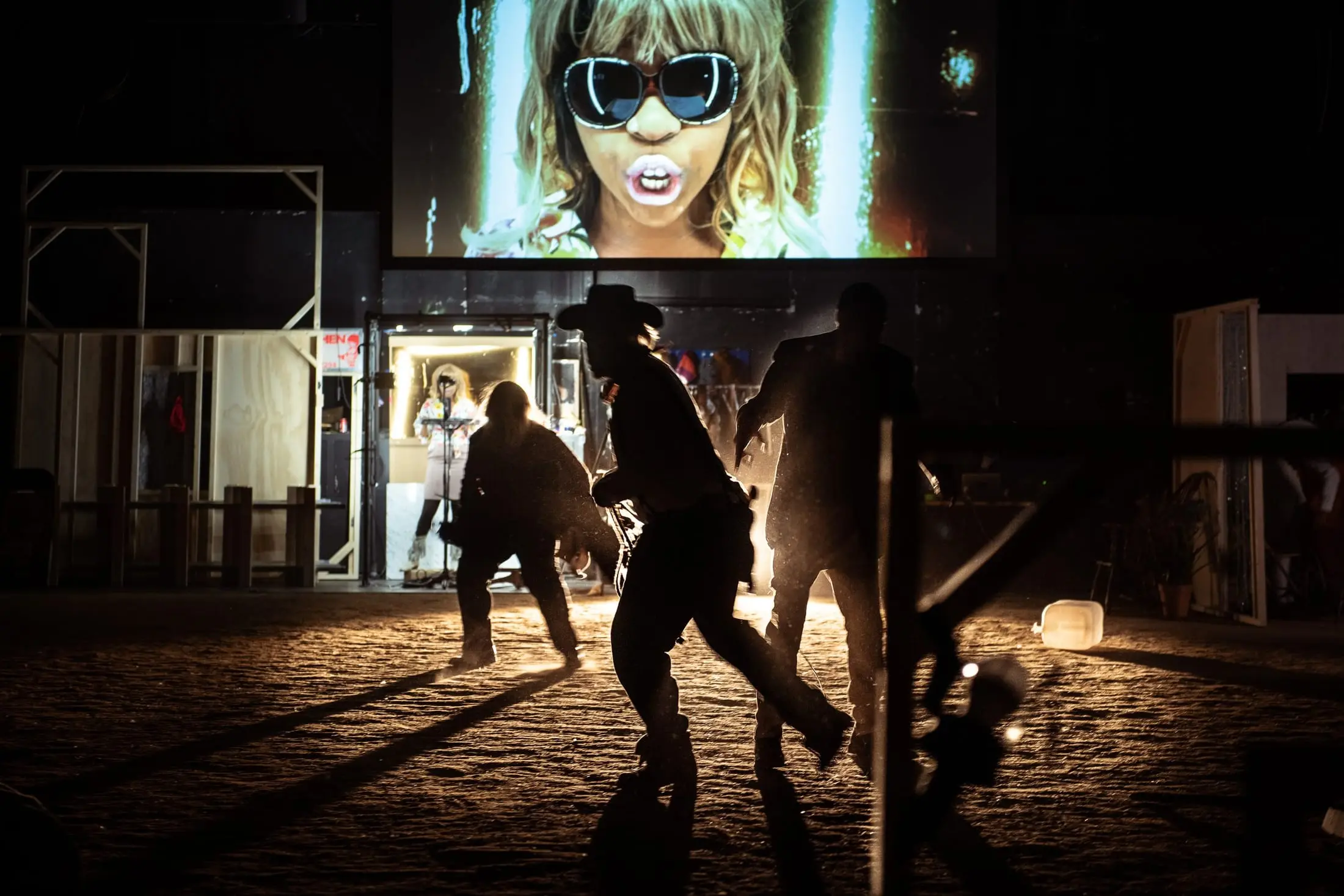 Dark Noon performers in silhouette on the dusty set, a projected face glowing above the town