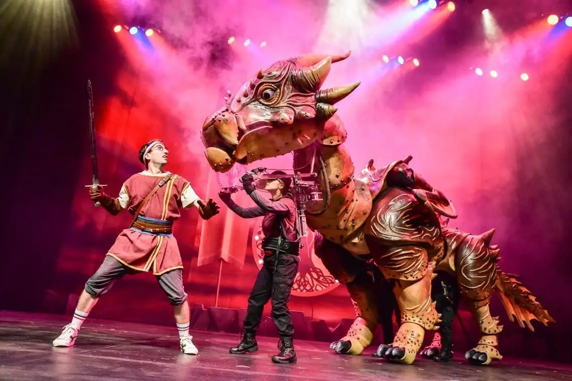 A hero with a sword faces a huge dragon puppet under pink stage light
