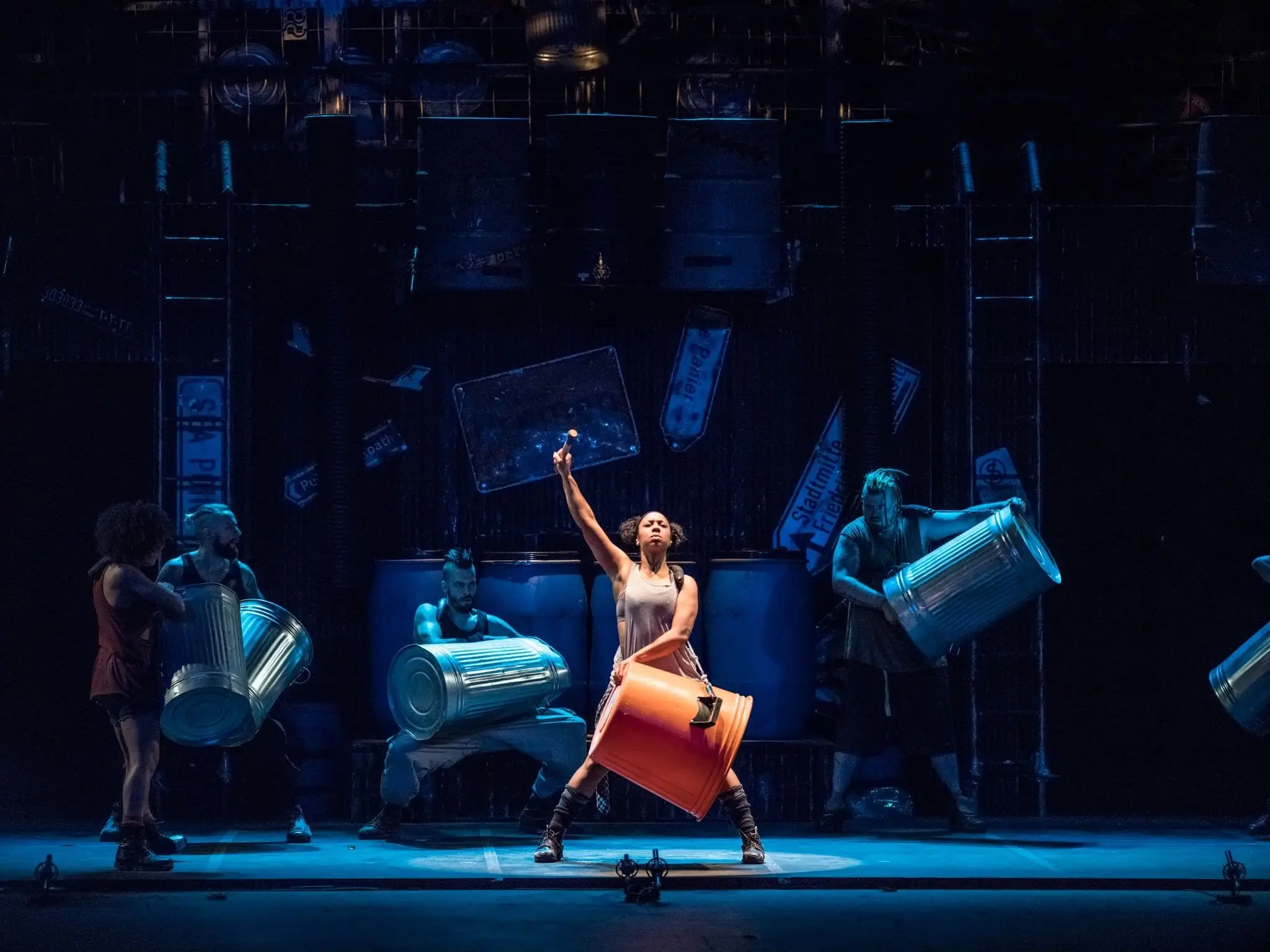 A performer strikes an oil drum, arm raised, in a pool of light