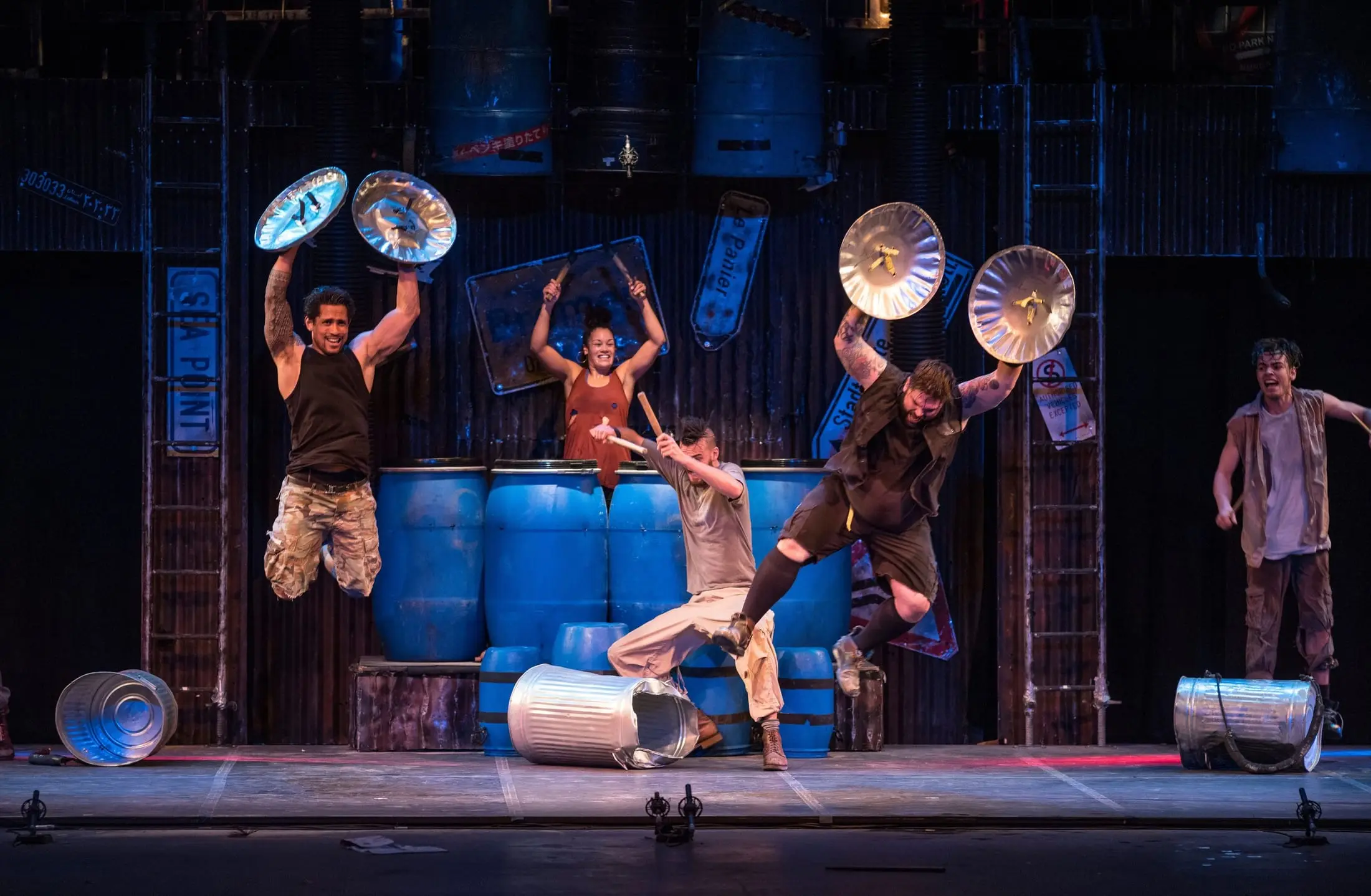 The STOMP company mid-leap on stage, oil drums and bin lids held high under blue light