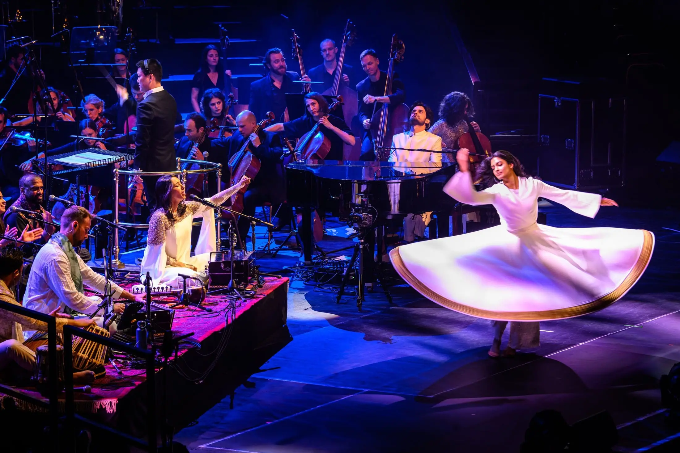A whirling dancer in white on stage with the orchestra and singers under deep blue light