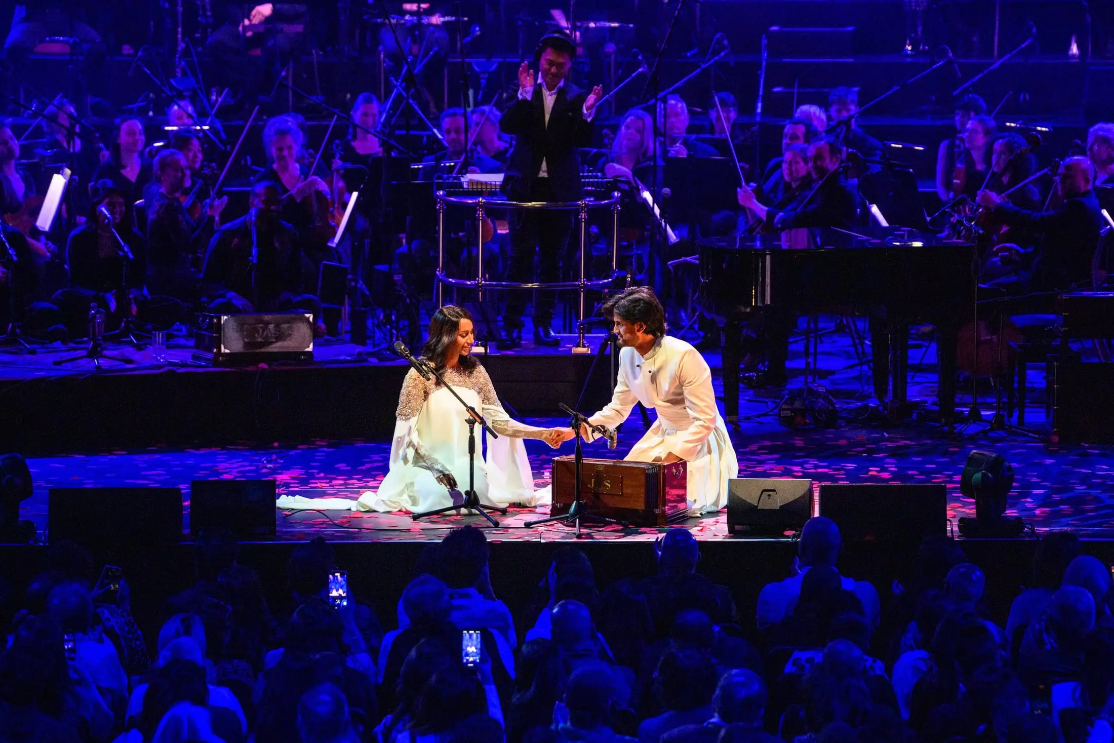Singer and harmonium player seated centre stage in front of the orchestra