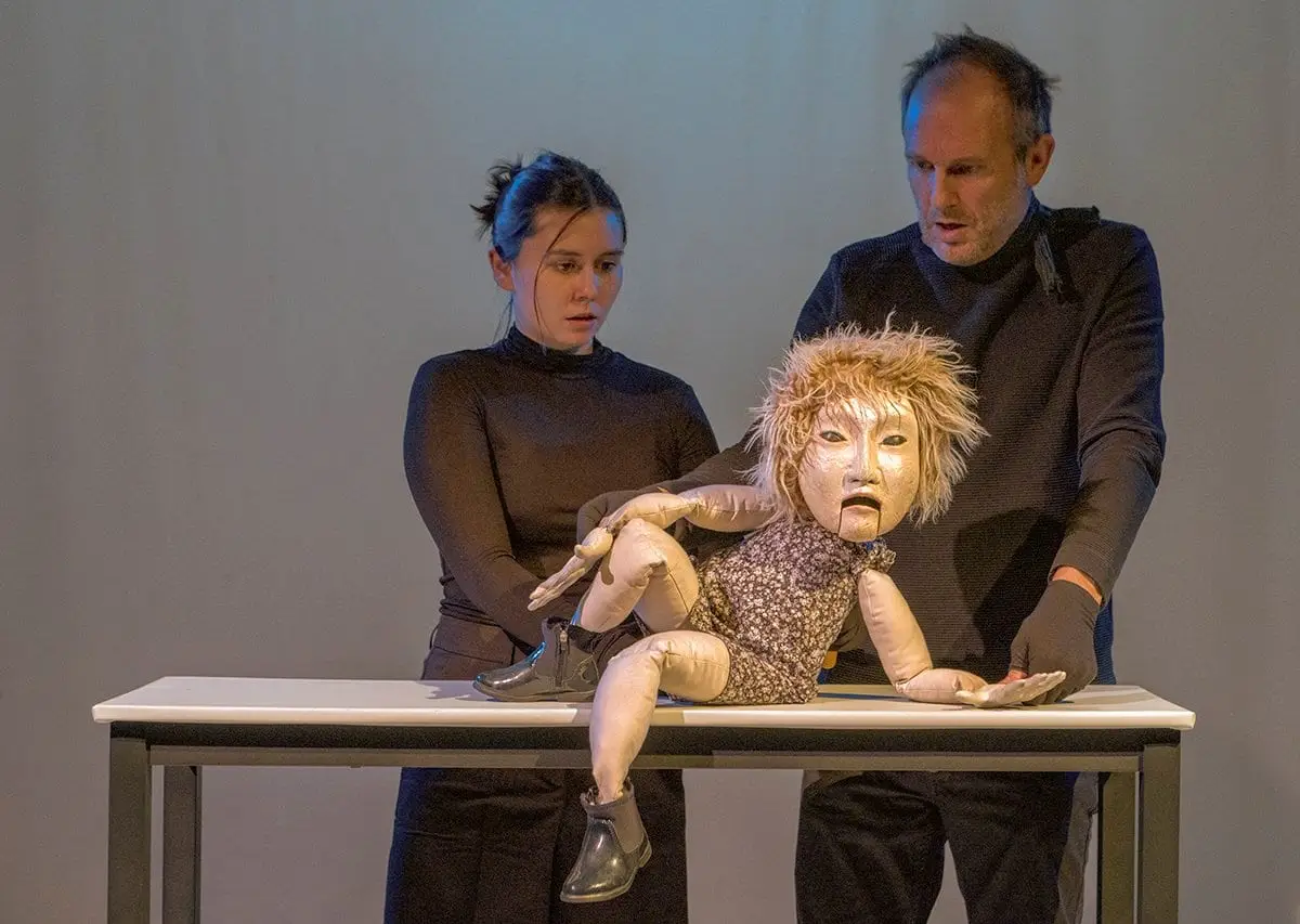 A puppet in a patterned dress leans on a table between two puppeteers