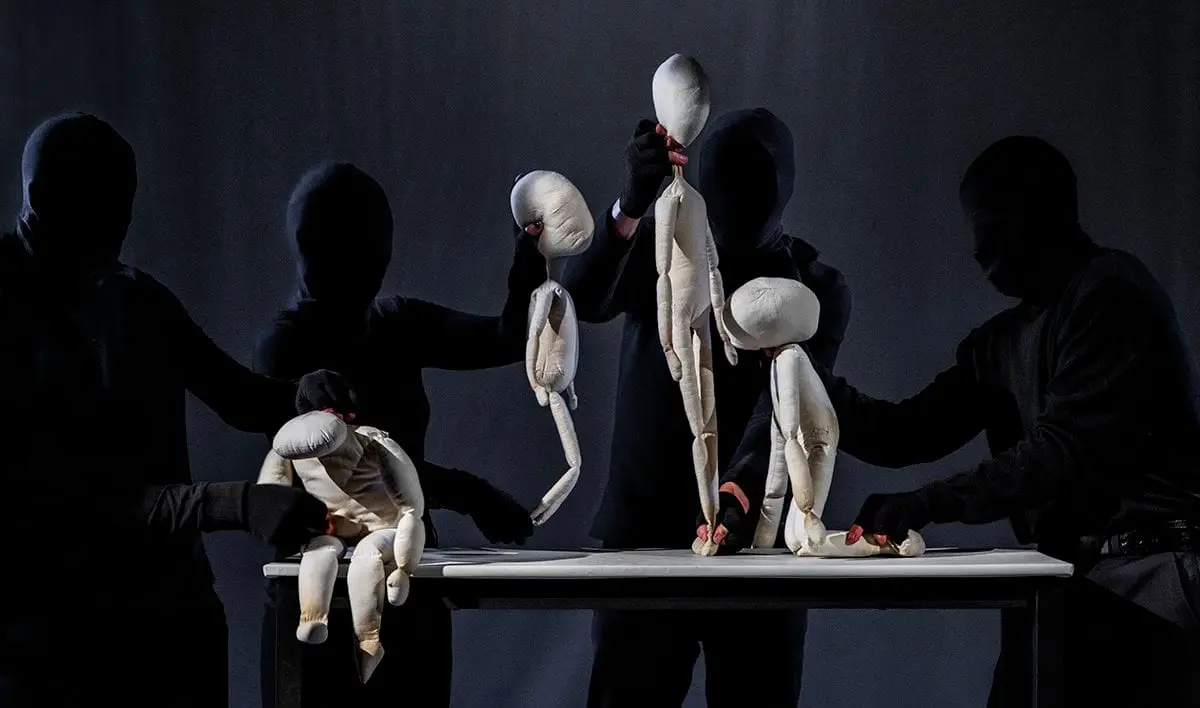 Two bare puppets embrace on a table, puppeteers in silhouette