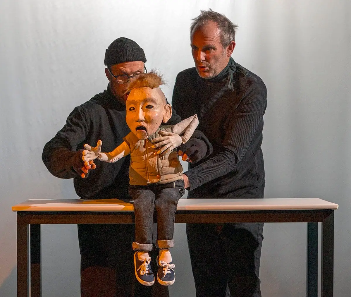 A puppet in a jumper sits on a table between two puppeteers