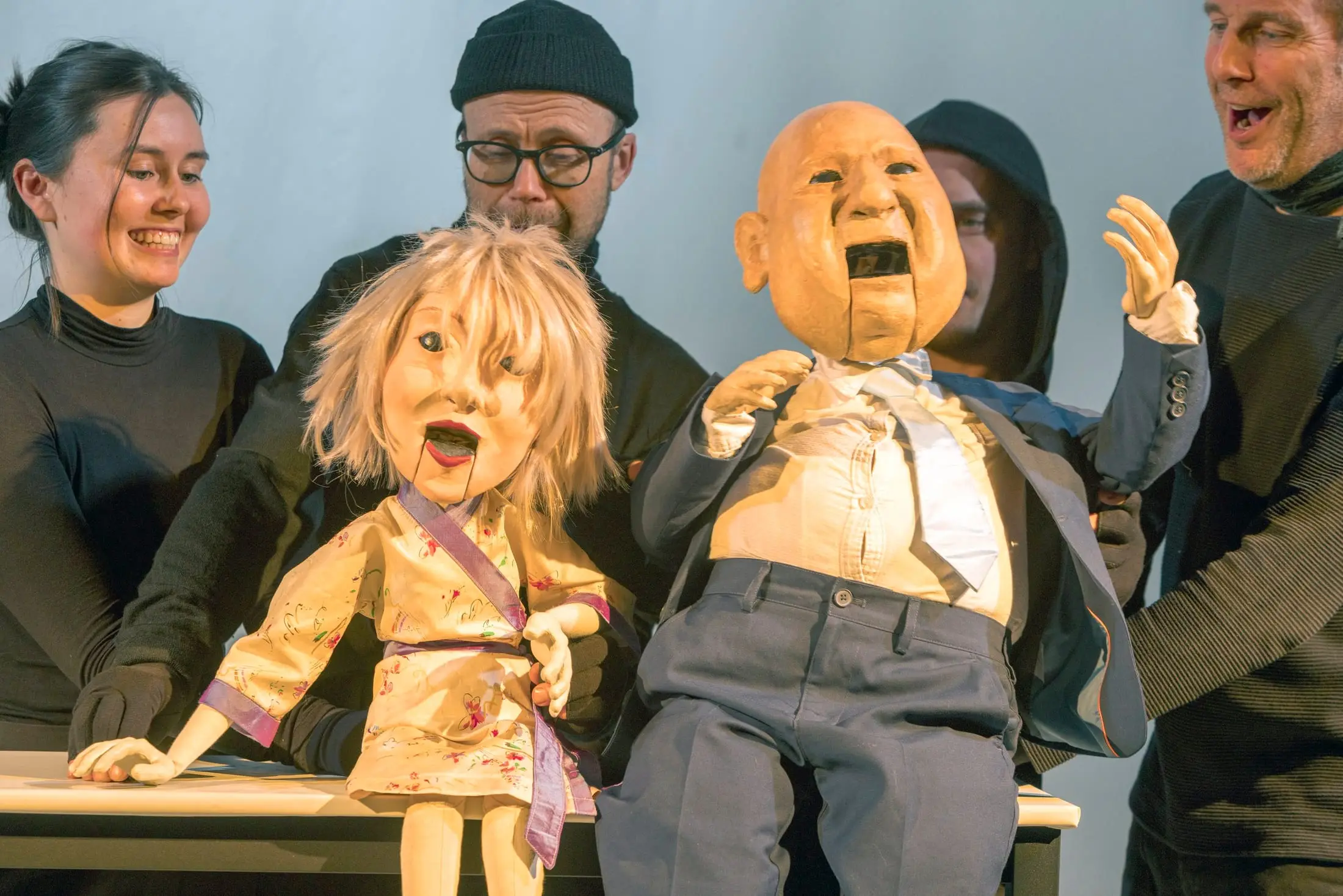 Two puppets in mid-conversation at a table, their puppeteers laughing behind them